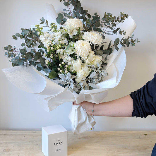 Wrapped bouquet of white and green flowers resting on a timber table beside a boxed soy candle
