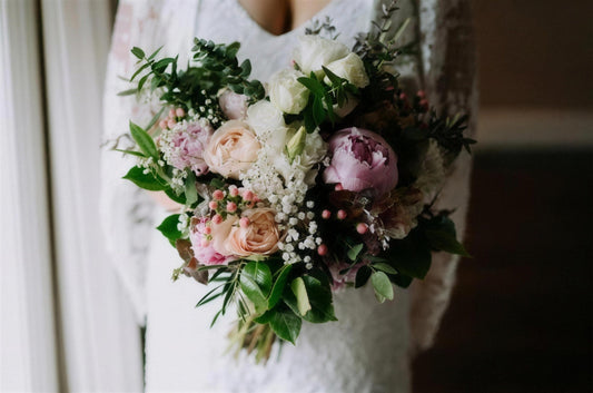A bride in soft window light holding a bouquet of peonies, garden roses and eucalyptus