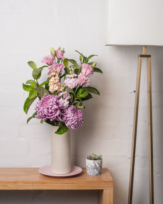 A bouquet of soft pink and purple flowers in a tall ceramic vase on a wooden console in a warm living room
