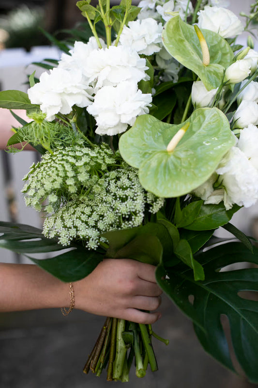 Hand holding a bouquet of white carnations, anthurium and monstera foliage against a softly blurred interior