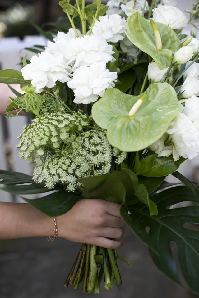 Hand holding a bouquet of white carnations, anthurium and monstera foliage against a softly blurred interior