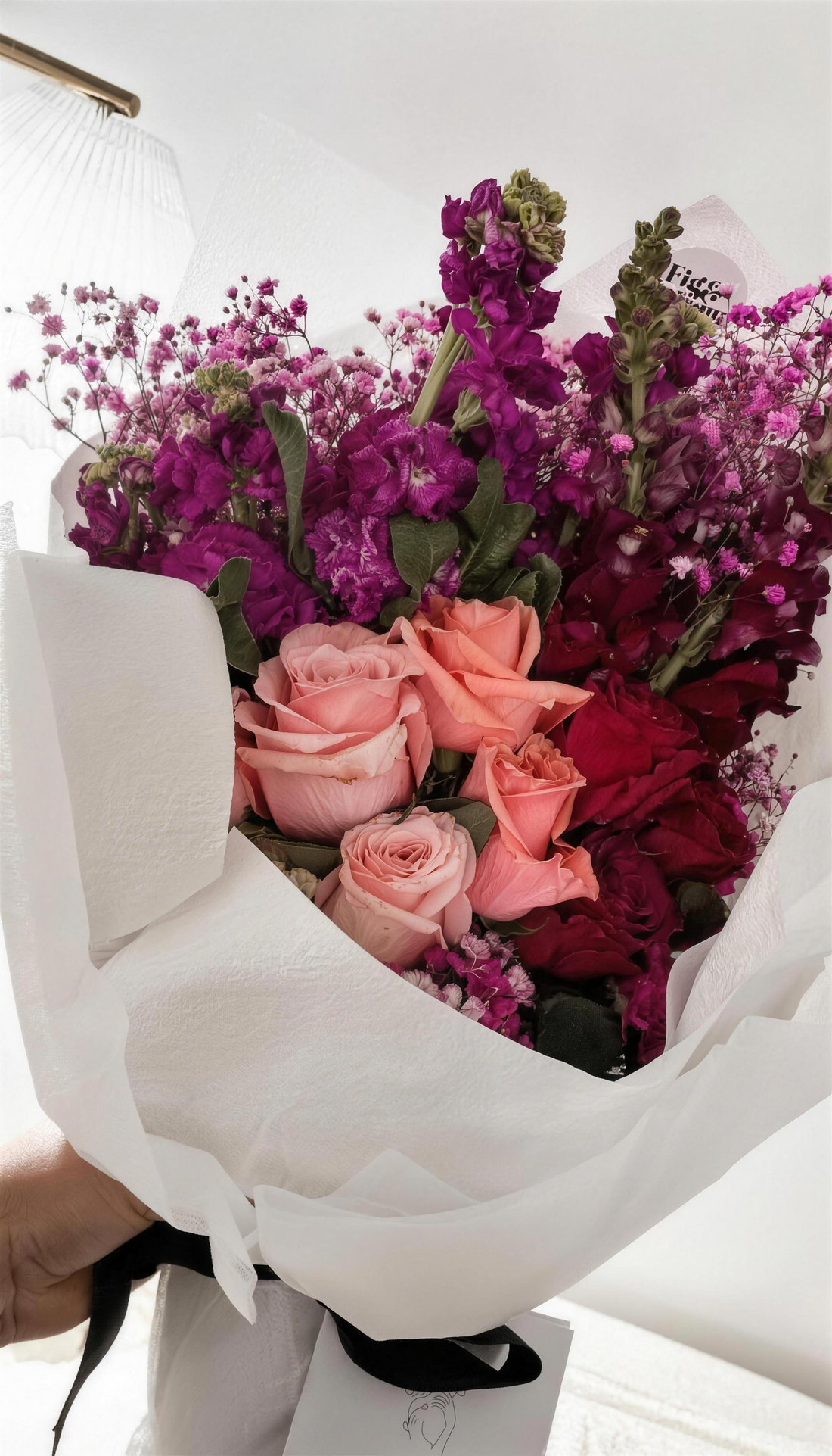 A hand holding a vibrant, considered bouquet — pink, peach, purple and deep red blooms