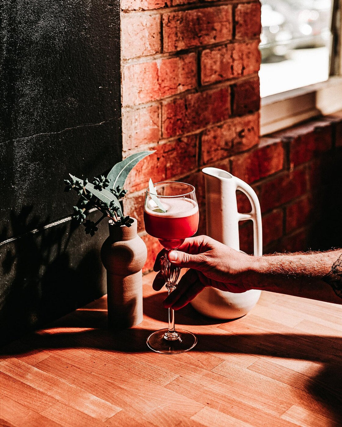 A hand holding a red cocktail beside a sunlit brick window