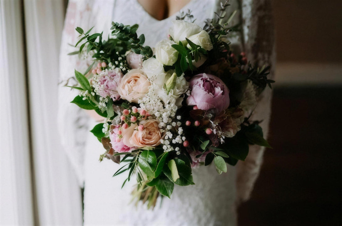 A bride in soft window light holding a bouquet of peonies, garden roses and eucalyptus