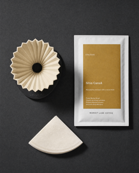 Flat lay of The Filter Ritual pour-over gift set on charcoal: Origami Air dripper and black AS holder seen from above, natural cone filters and a 250g bag of Market Lane Sitio Canaa filter coffee.