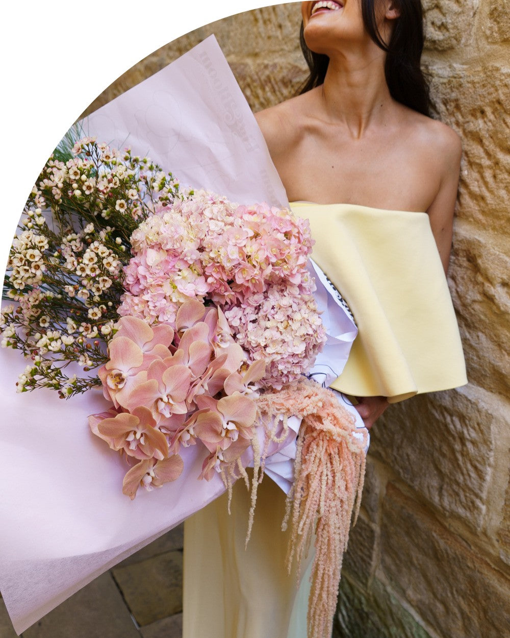 A woman holds the Carmen arrangement from the High Floral Design collection, pink hydrangea and blush phalaenopsis orchids with trailing amaranthus, wrapped in lilac paper against a sandstone wall