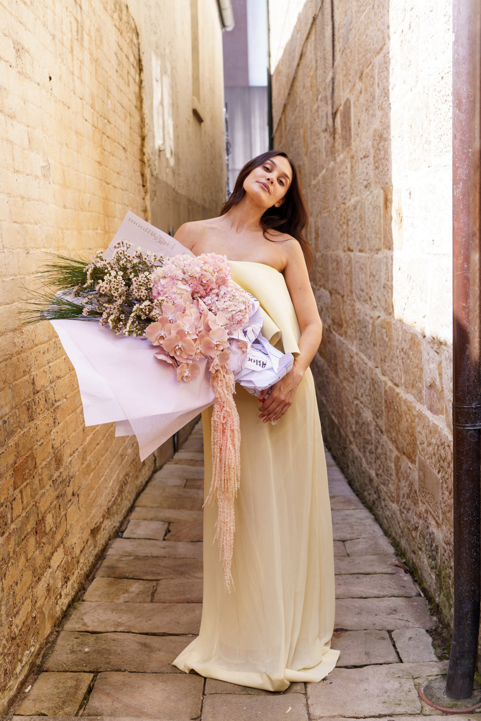 Full-length shot of a model in a pale yellow gown carrying the Carmen arrangement down a laneway