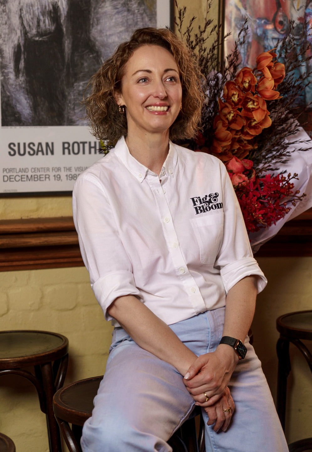 Kellie Brown seated with hands clasped, in a white Fig & Bloom shirt and pale jeans, in front of framed posters