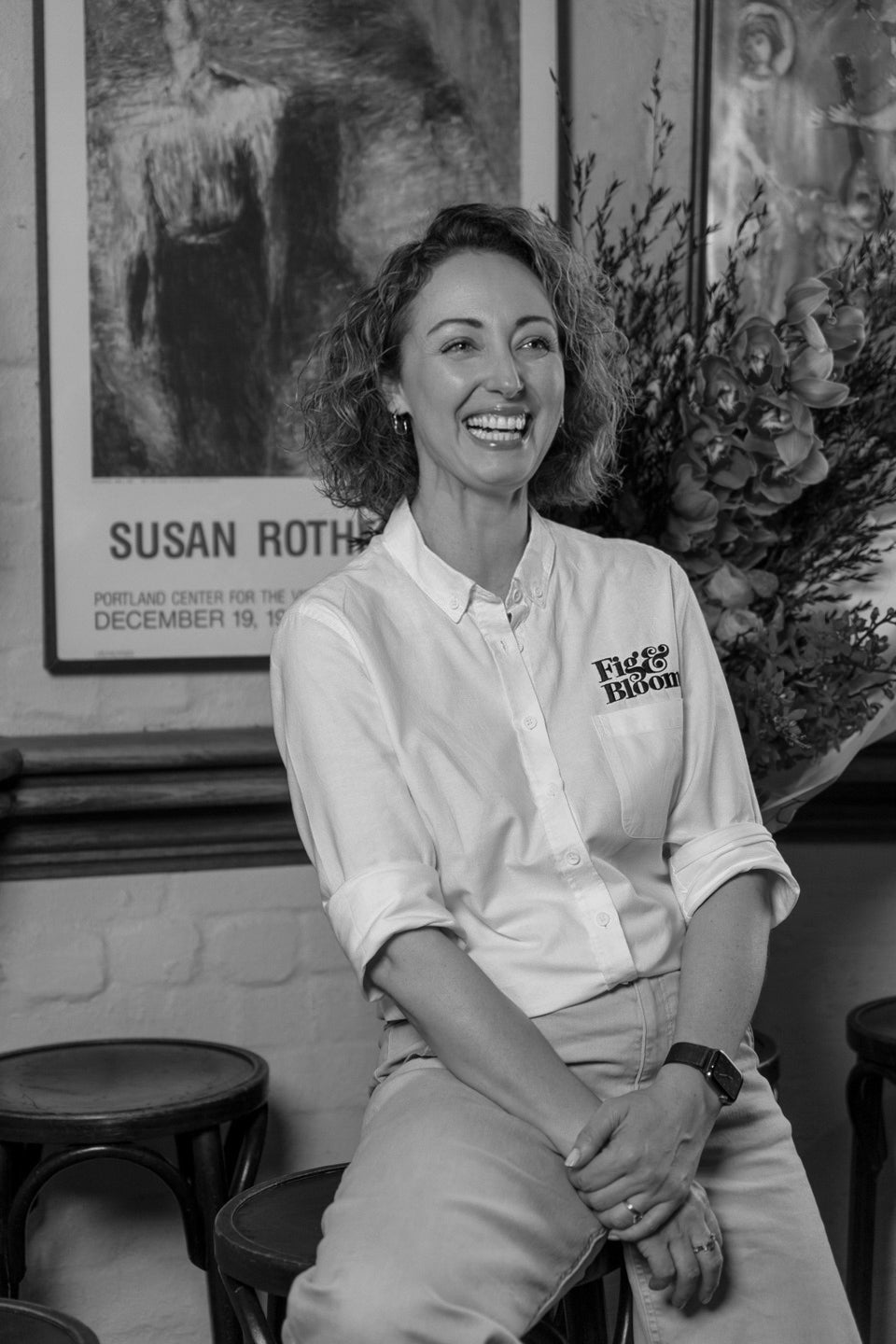 Black and white portrait of Kellie Brown seated on a bar stool, smiling and looking off camera