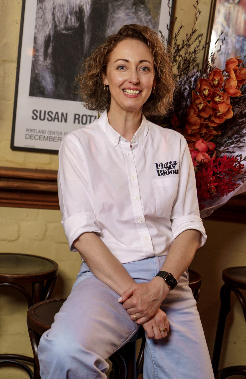 Kellie Brown, co-founder of Fig & Bloom, seated on a bar stool in a white Fig & Bloom shirt, smiling, with orange orchids behind her