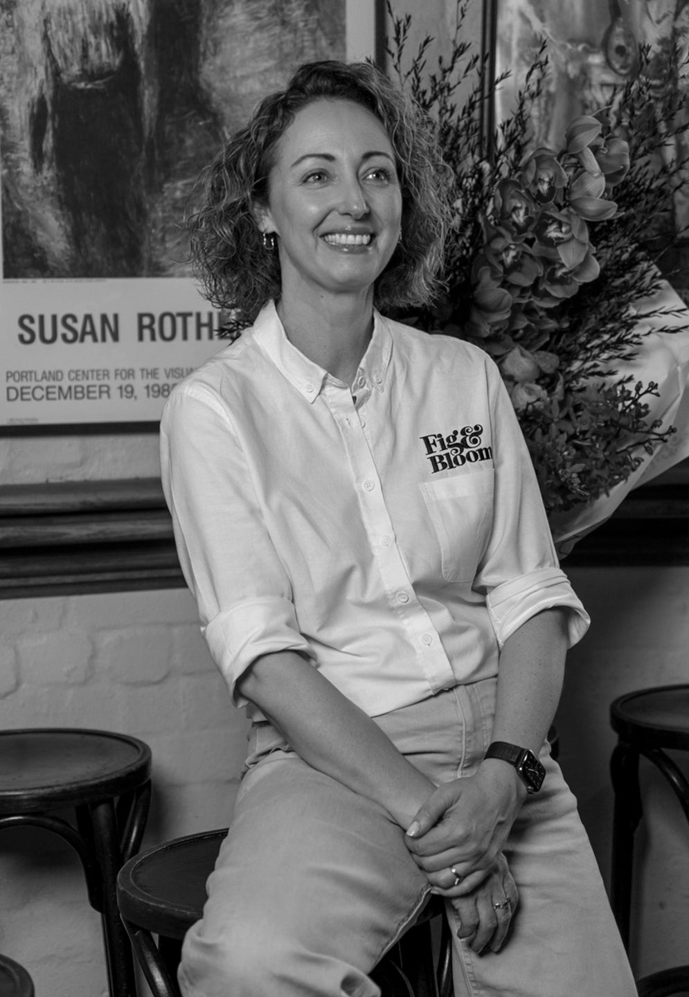 Black and white portrait of Kellie Brown laughing, seated in front of framed posters