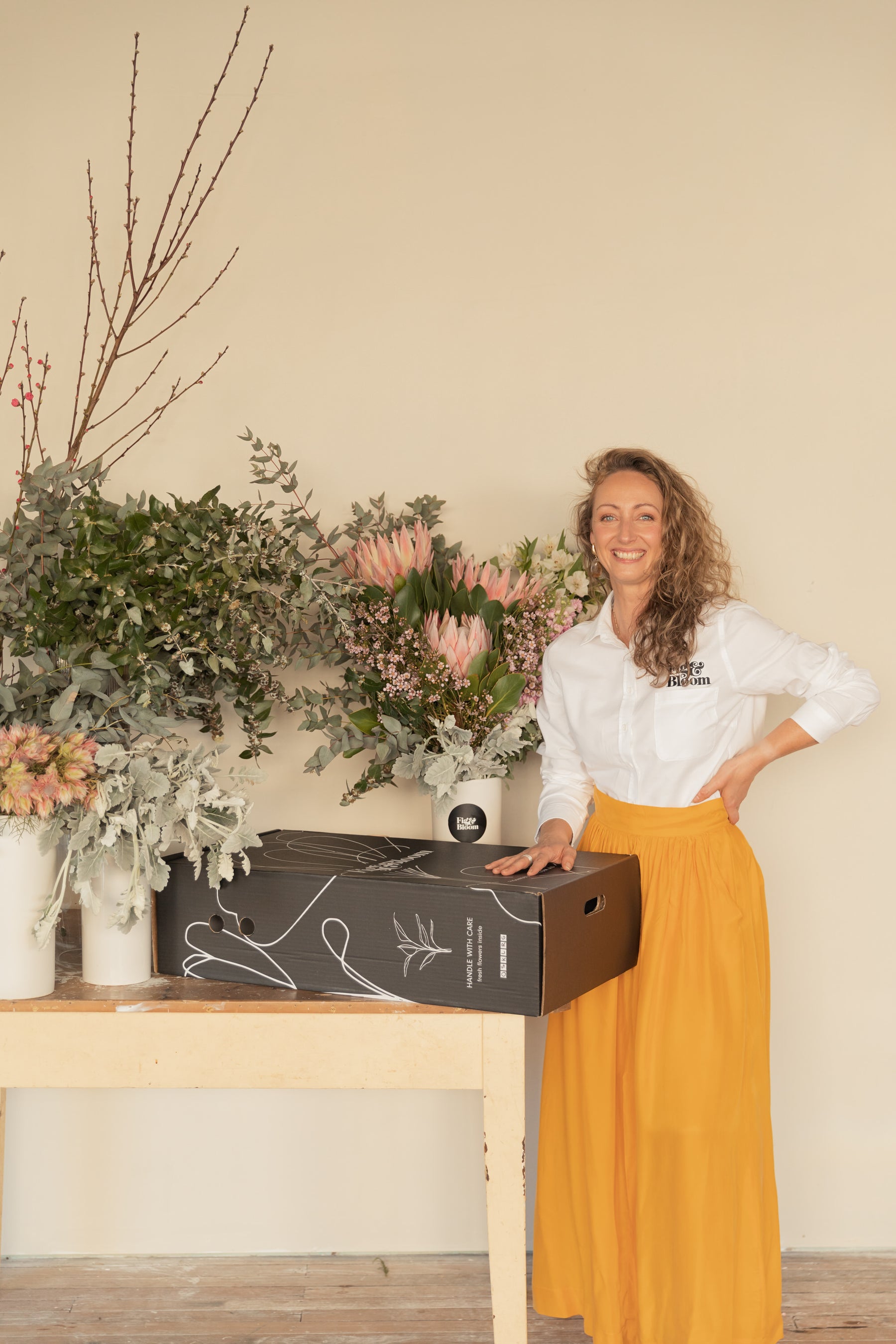 Kellie Brown smiling beside a Fig & Bloom flower box, with vases of proteas and eucalyptus behind her