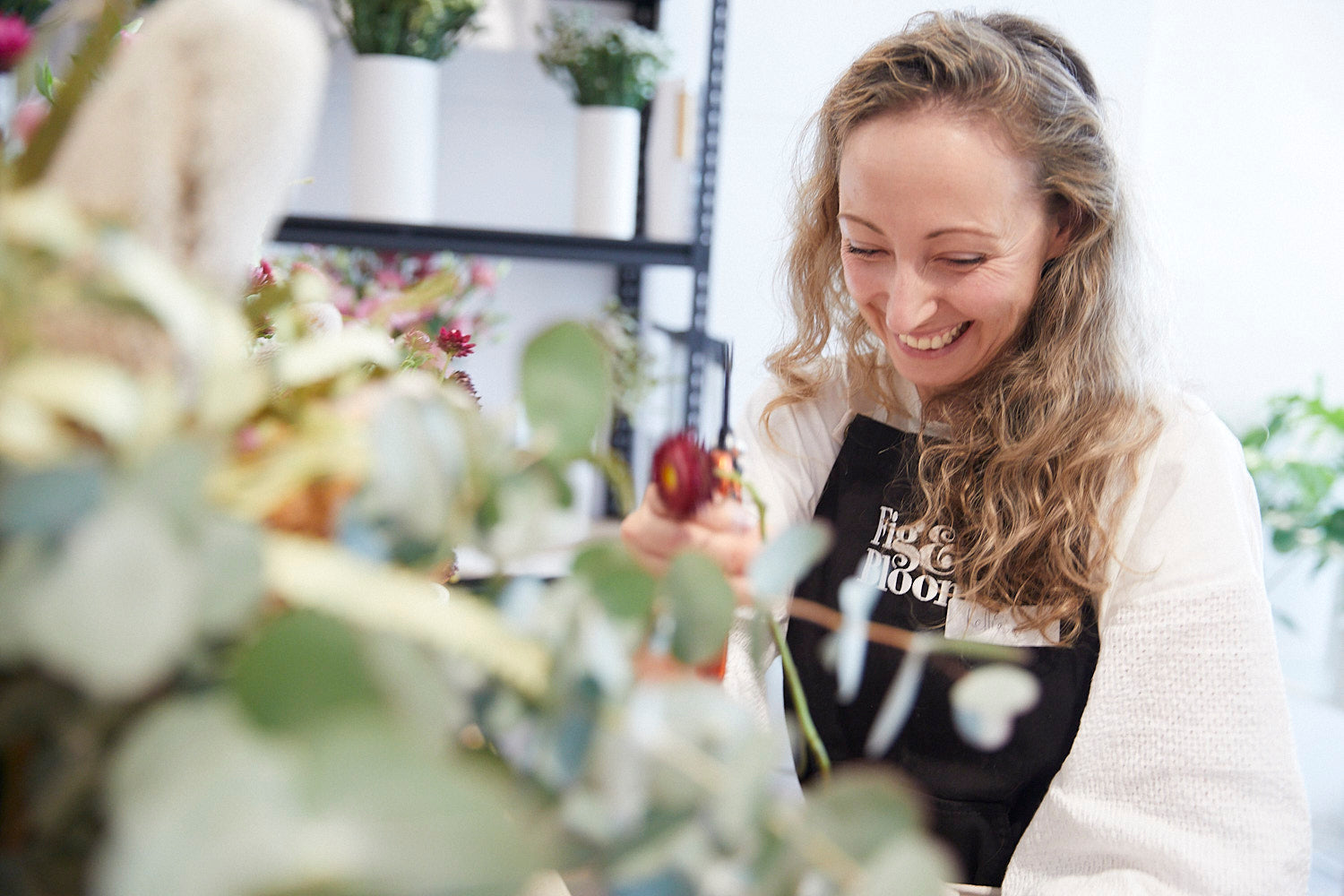 Kellie Brown smiling as she arranges flowers, seen through foreground eucalyptus