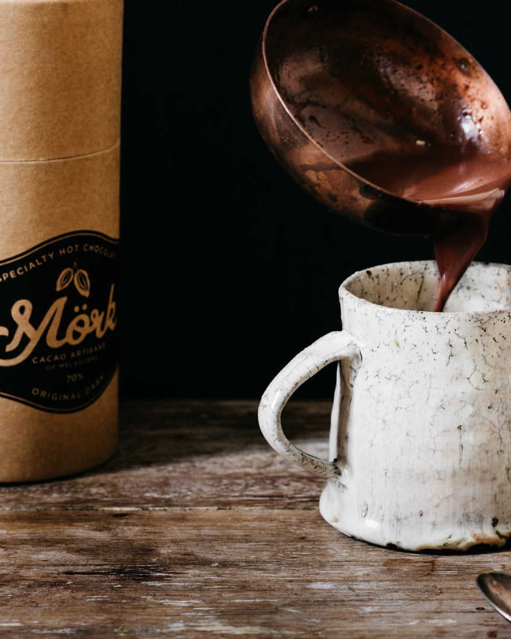Mörk hot chocolate being poured from a copper ladle into a ceramic mug beside the 250g tube