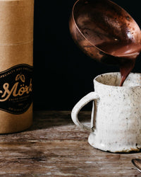 Mörk hot chocolate being poured from a copper ladle into a ceramic mug beside the 250g tube