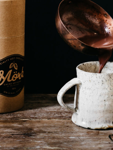 Mörk hot chocolate being poured from a copper ladle into a ceramic mug beside the 250g tube