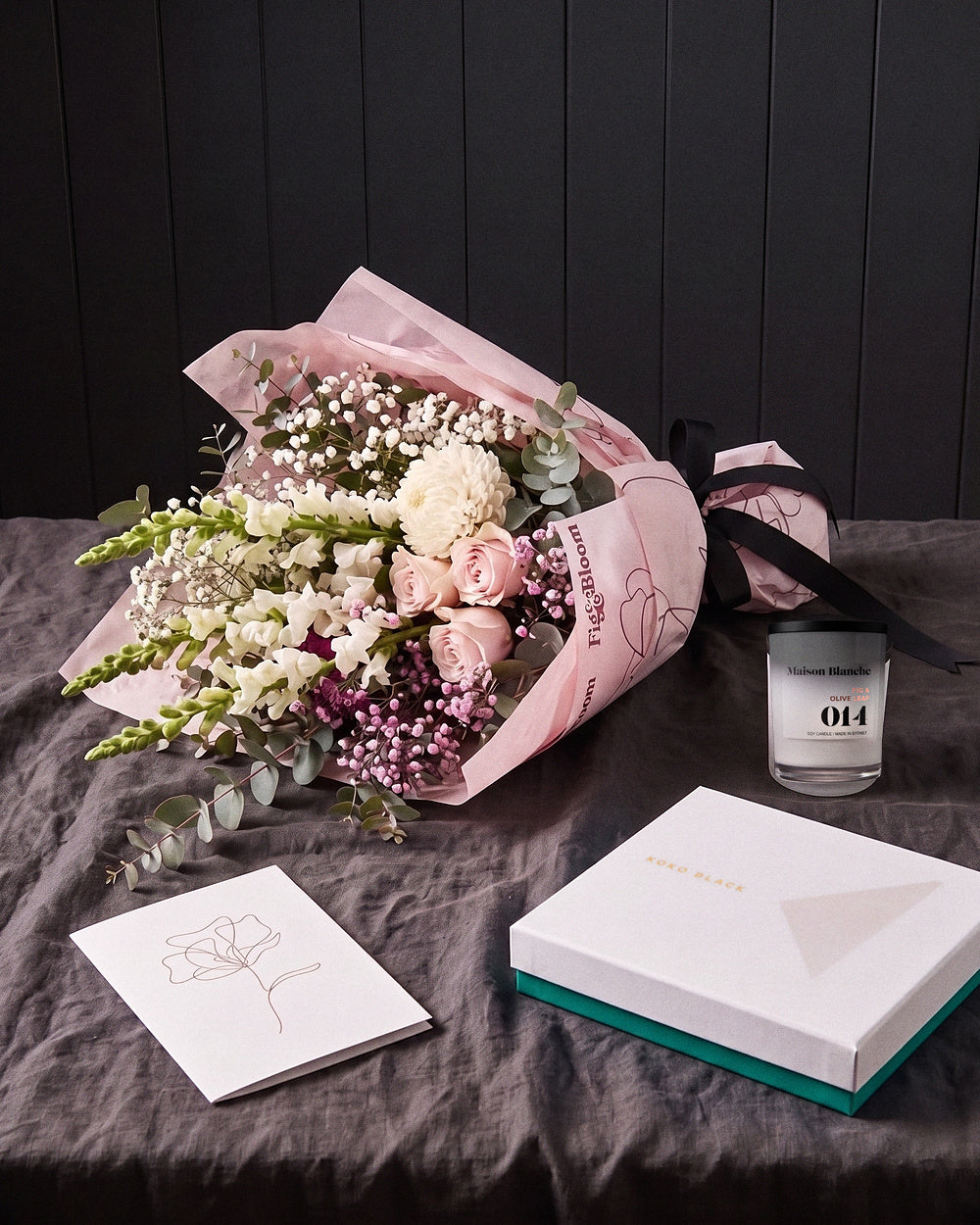 The Fuss resting on charcoal linen in window light, with the candle, chocolates and greeting card alongside.