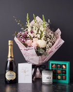 The Lot, the Osaka bouquet tied with black ribbon on dark stone, with House of Arras Brut Elite, the Koko Black nine-piece selection, the Fig & Olive Leaf candle and the greeting card.
