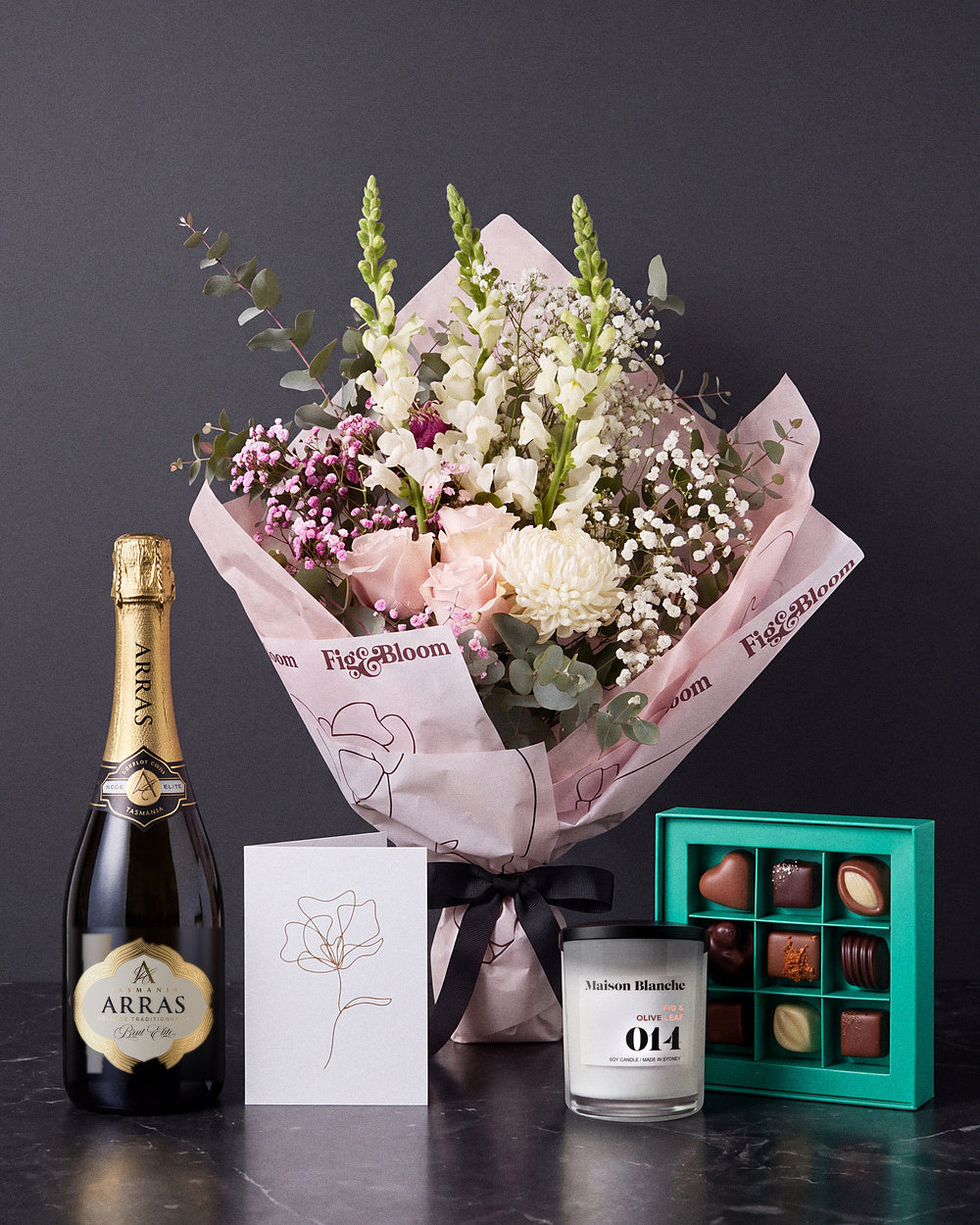 The Lot, the Osaka bouquet tied with black ribbon on dark stone, with House of Arras Brut Elite, the Koko Black nine-piece selection, the Fig & Olive Leaf candle and the greeting card.