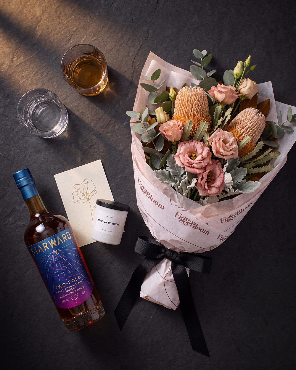 The Nightcap photographed from above on charcoal slate: the Broome Gesture bouquet, Starward Two-Fold, two whisky tumblers, the candle and the greeting card.