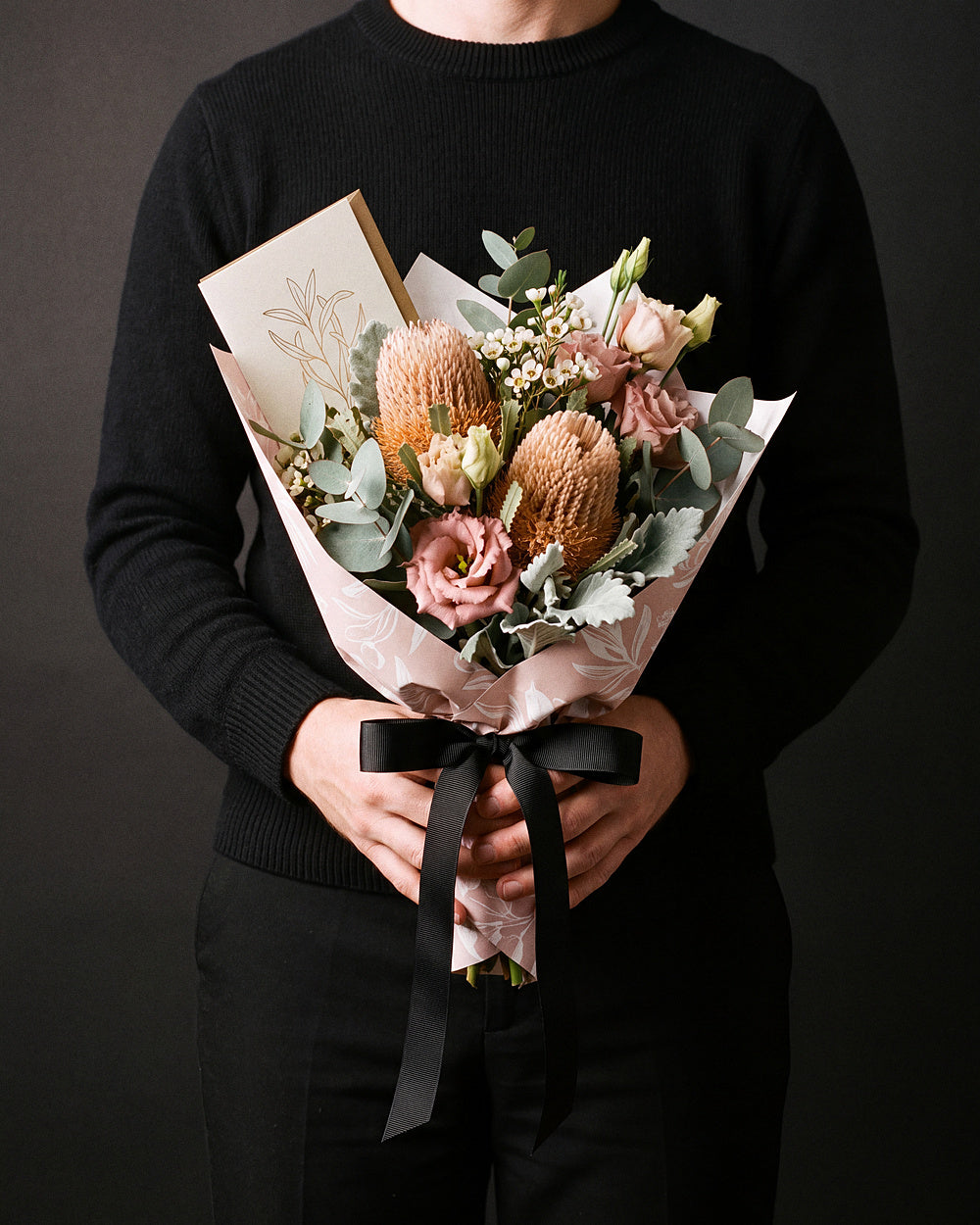 The Nightcap, the Broome Gesture bouquet held at chest height with the greeting card tucked into the wrap, against a charcoal backdrop.