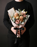 The Nightcap, the Broome Gesture bouquet held at chest height with the greeting card tucked into the wrap, against a charcoal backdrop.