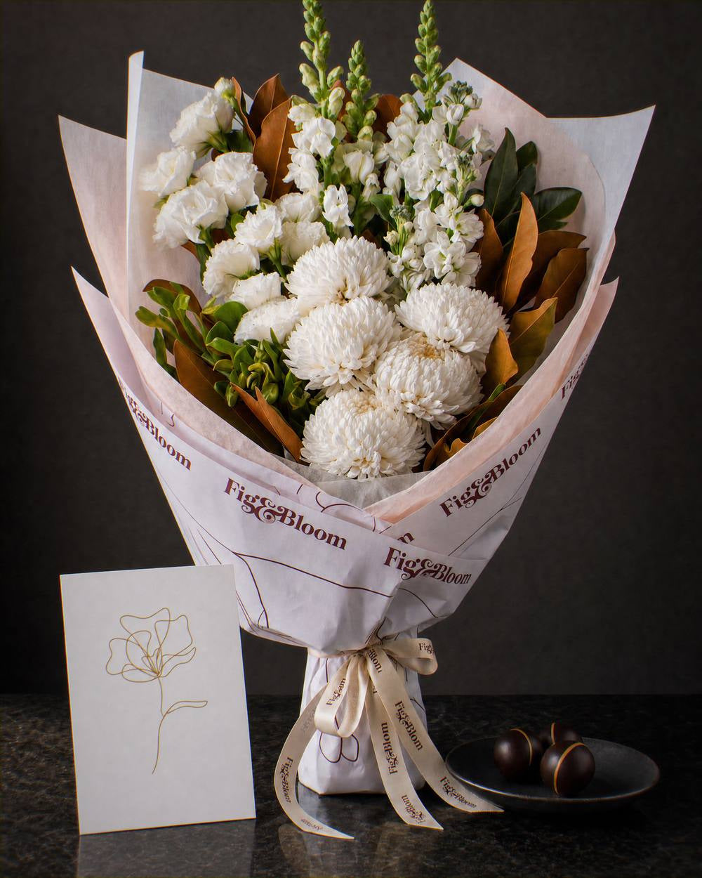 The Toast, an all-white Pyrenees bouquet with a greeting card and a plate of dark chocolate marbles, on dark stone.
