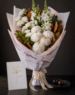 The Toast, an all-white Pyrenees bouquet with a greeting card and a plate of dark chocolate marbles, on dark stone.
