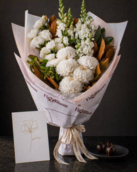 The Toast, an all-white Pyrenees bouquet with a greeting card and a plate of dark chocolate marbles, on dark stone.