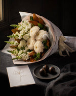 The Toast, the Pyrenees bouquet resting on charcoal linen in window light, with the greeting card and dark chocolate marbles.