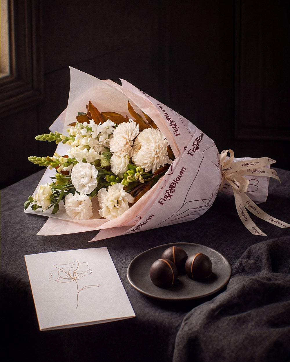 The Toast, the Pyrenees Gesture bouquet resting on charcoal linen in window light, with the greeting card and dark chocolate marbles.