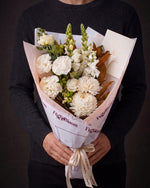 The Toast, the Pyrenees Gesture bouquet held at chest height with the greeting card tucked into the wrap, against a charcoal backdrop.