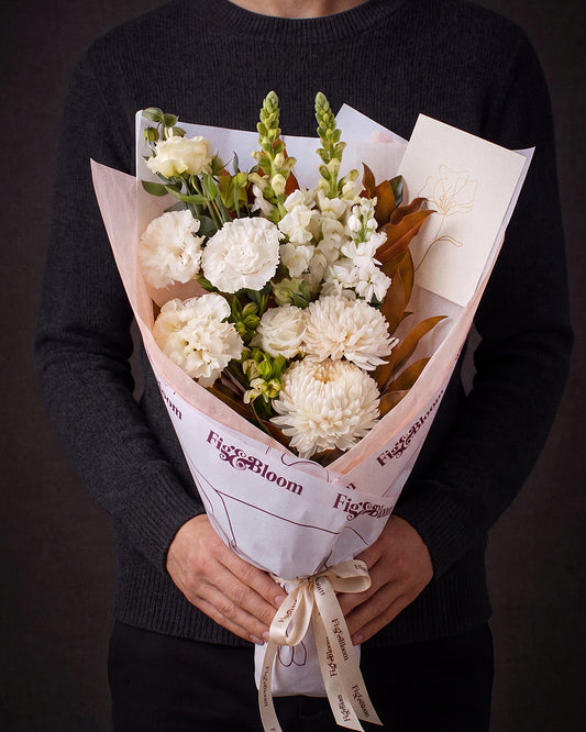 The Toast, the Pyrenees Gesture bouquet held at chest height with the greeting card tucked into the wrap, against a charcoal backdrop.
